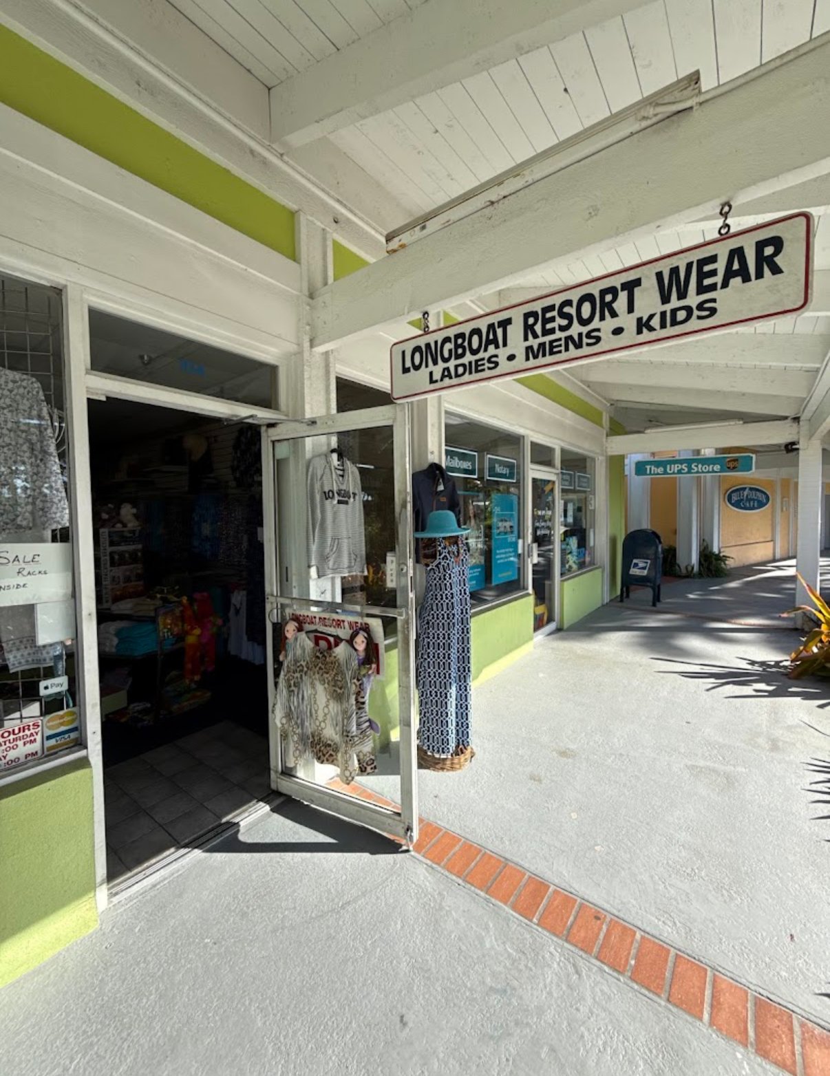 The entrance to Longboat Resort Wear at The Centre Shops, with the Ladies · Mens · Kids sign over the door, a sundress and hooded top on display, and the breezy plaza walkway alongside